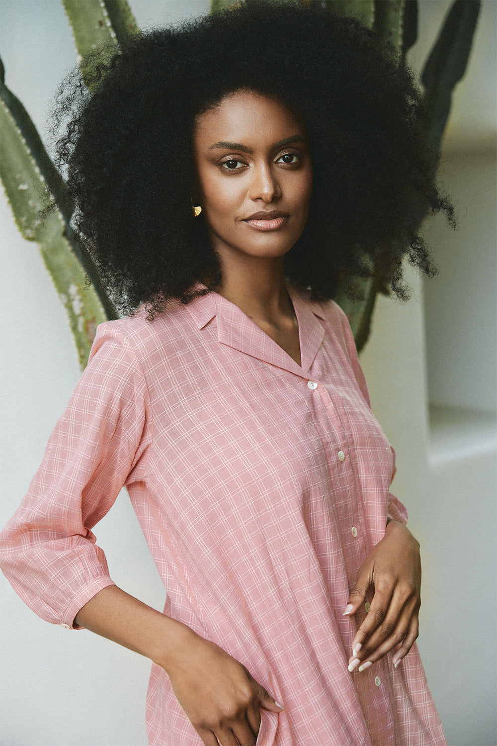 Model wearing the Blush Mist Dress in soft pink checks, with a relaxed collar and side seam detail, standing beside a cactus plant.