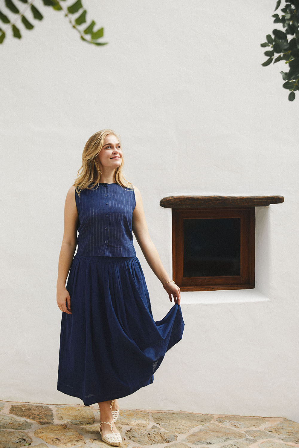 Model standing against a white wall with a small wooden window, wearing Indigo Dream Set featuring a sleeveless indigo top with white pinstripes and a matching mid-calf skirt.