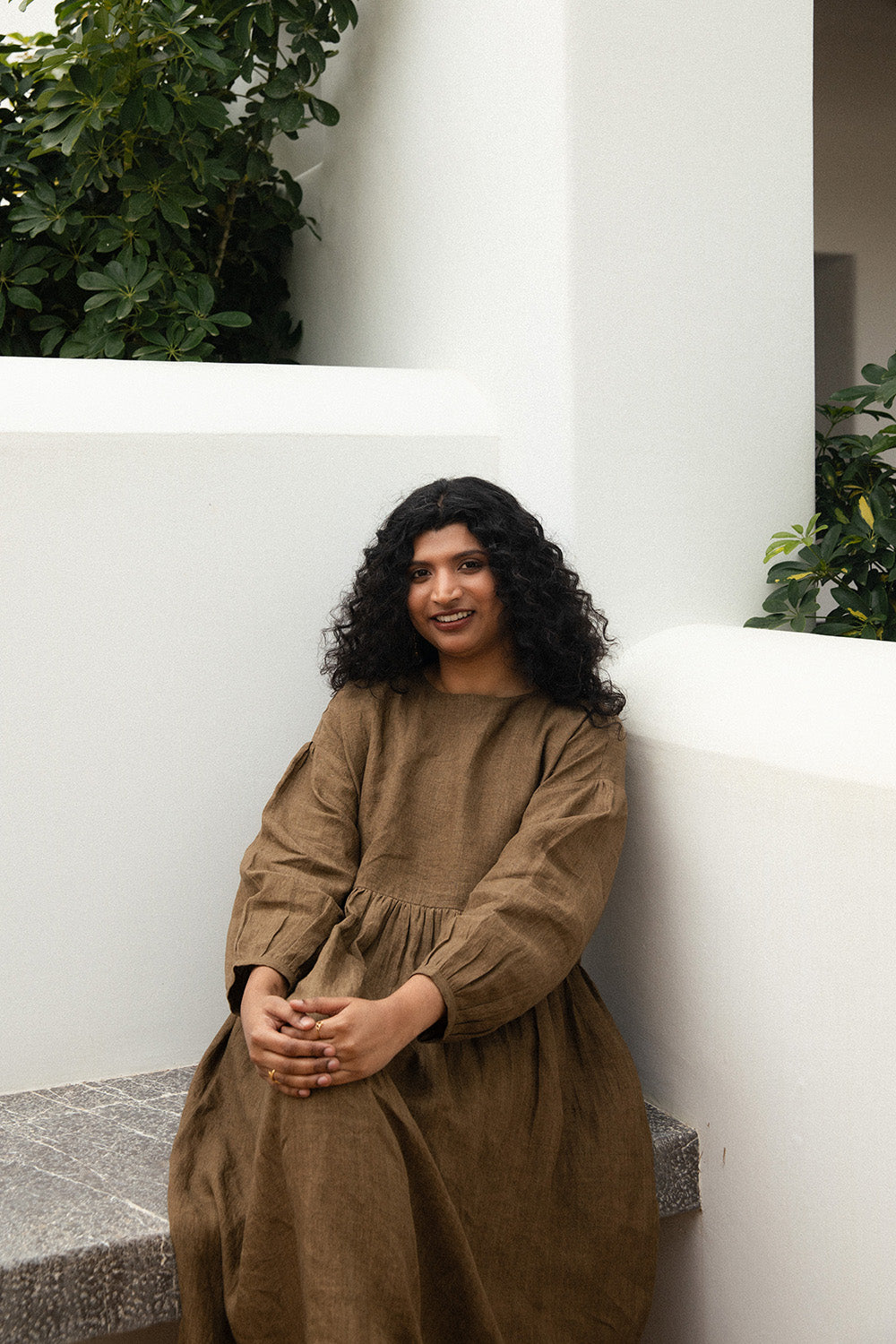 Apoorva M Sivakumar, founder of Meenal, seated and smiling while wearing the Olive Drift Dress in a minimalist courtyard setting.
