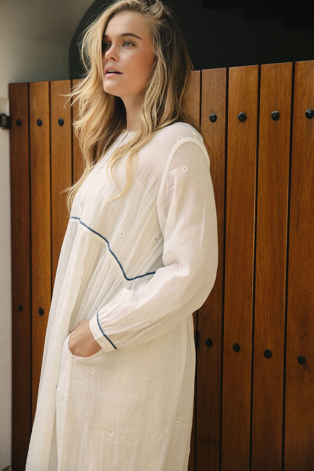 Model wearing the Morning Dew Dress in white with blue piping, standing in front of a wooden door with her hand in the pocket.