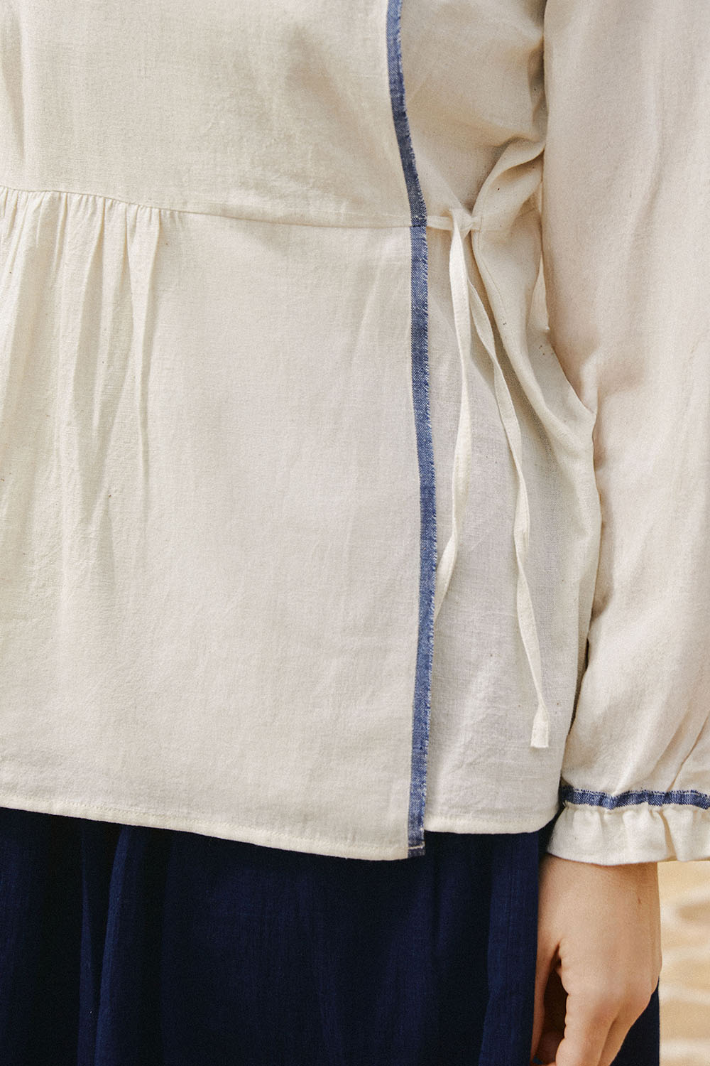 A close-up of Morning Fold Wrap Top in off-white with a soft round neckline, gathered waist, and indigo detailing on the sleeves and hem.