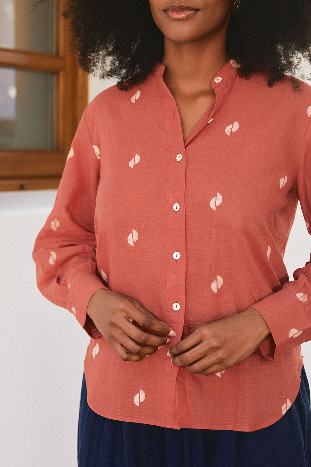 Close-up of model in the Sienna Moon Shirt in rust tone with subtle motifs, paired with a navy skirt, standing by a white wall.