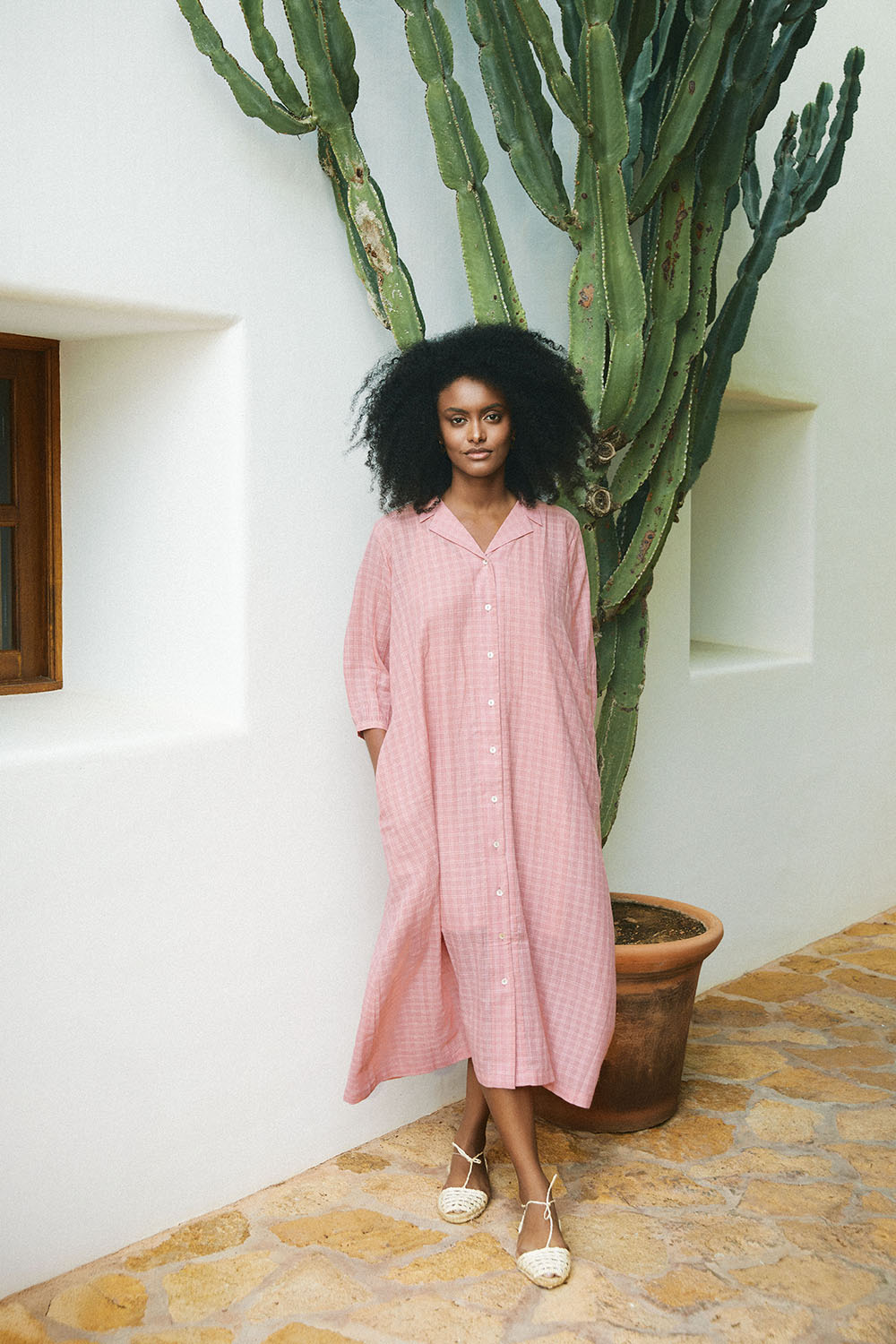 Model wearing the Blush Mist Dress in a soft pink check, standing beside a white wall and a cactus plant.