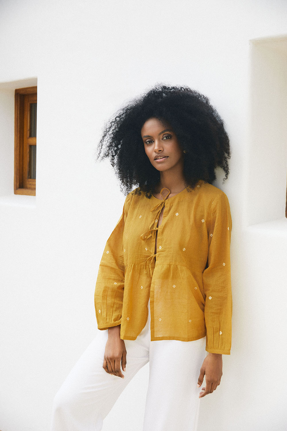 Model in the Golden Hues Front Tie Top in mustard yellow, styled with white pants, standing against a white wall.