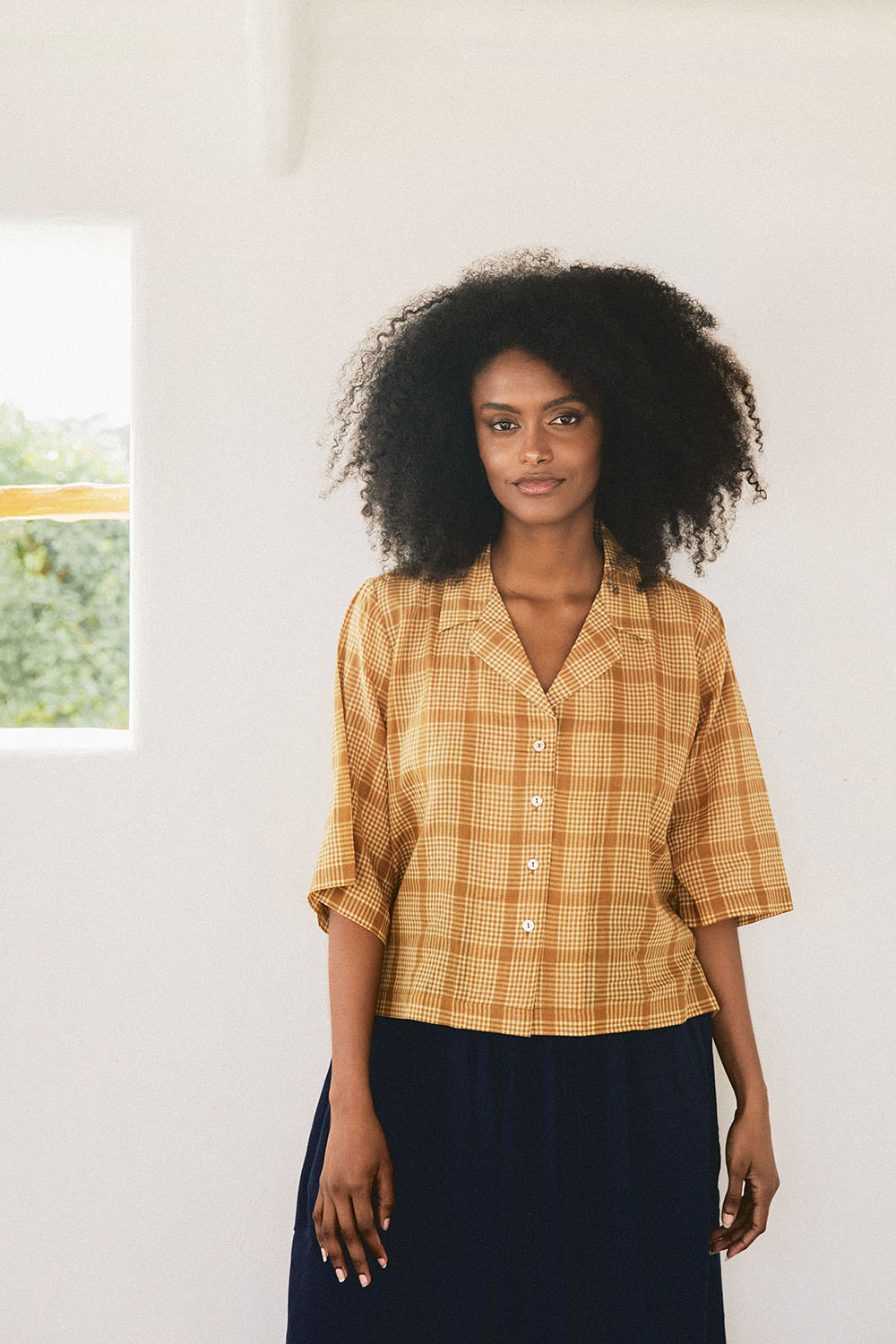 Model wearing the Golden Meadow Shirt in a warm mustard check, with a collar, buttoned front, and half sleeves, standing by a window-lit white wall.