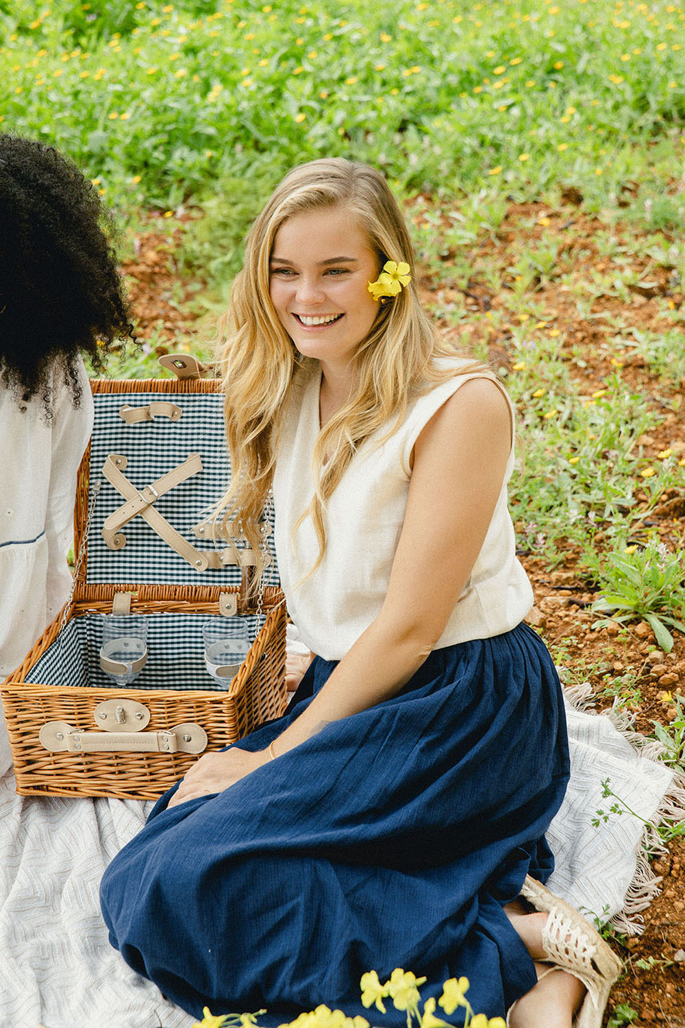 Two models seated outdoors on a picnic blanket; one wearing the Grain Breeze Vest Top in ivory with the Indigo Dream Skirt, beside a wicker picnic basket.