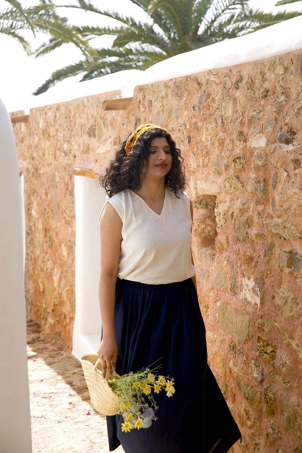 Apoorva, the founder, wearing the Grain Breeze Vest Top with the Indigo Dream Skirt, walking beside a sunlit stone wall while holding a straw bag filled with yellow wildflowers.