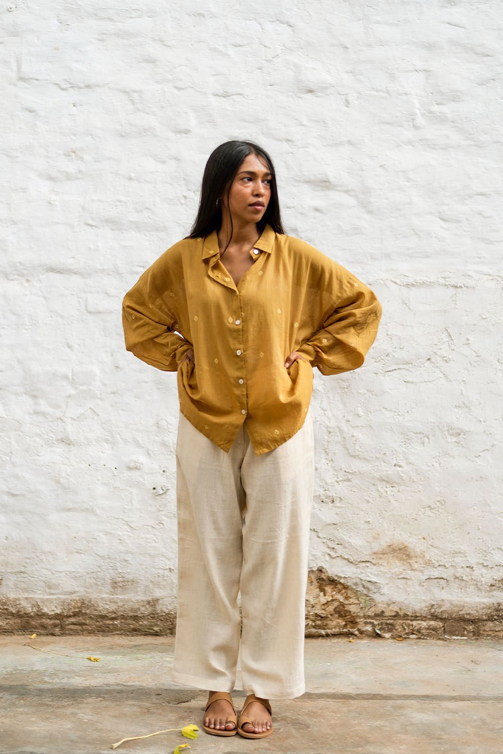 Model wearing Grain Pants in beige, styled with a mustard yellow shirt, standing against a white brick wall.