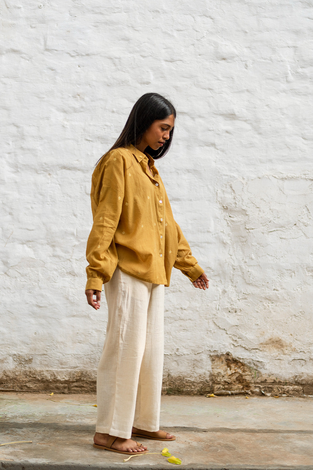 Model wearing Grain Pants in beige, styled with a mustard yellow shirt, standing against a white brick wall.