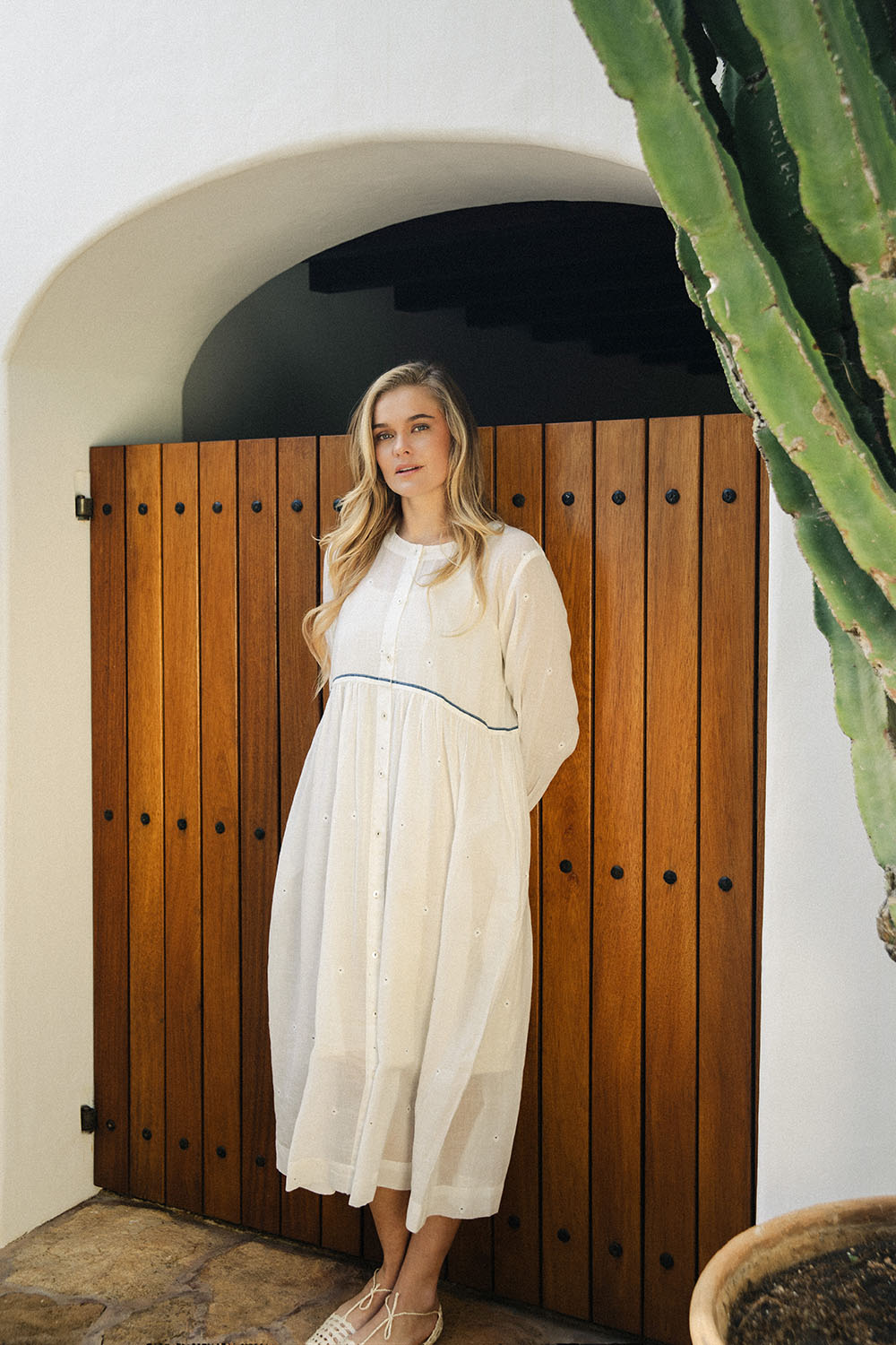 Model wearing the Morning Dew Dress in white, standing in front of a wooden arched door.