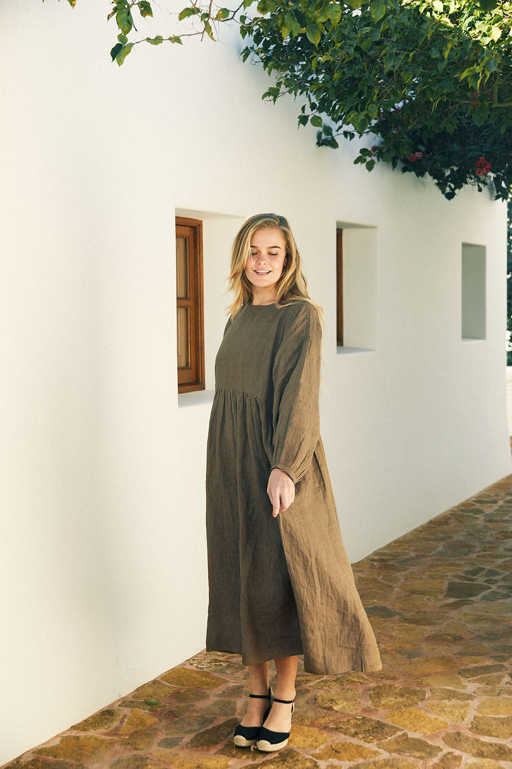 Model wearing the Olive Drift Dress in earthy olive green, standing beside a sunlit stone pathway next to a white wall.