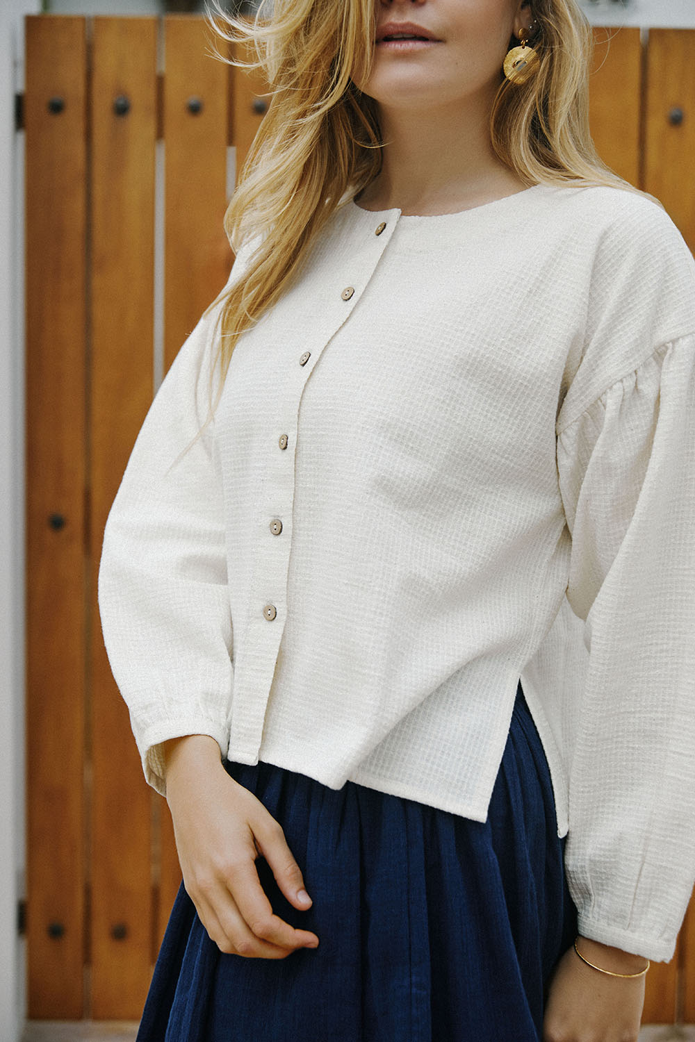 Close-up of model in the Pure Weave Blouse in ivory with long sleeves, round neckline, and front button closure, paired with Indigo Dream Skirt, posing in front of a wooden door.