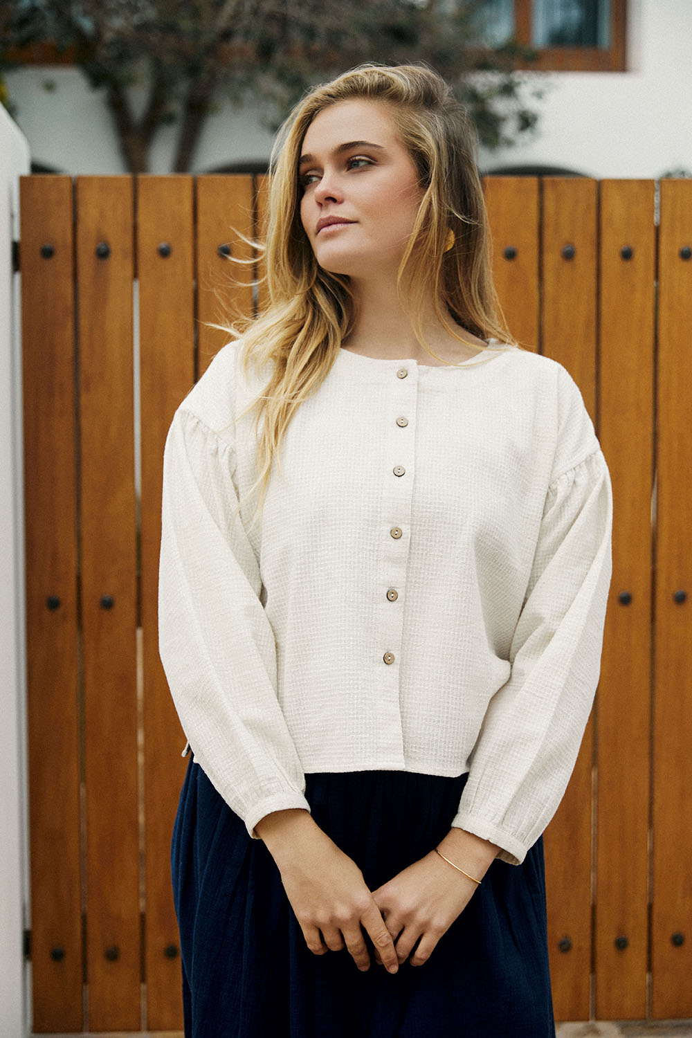 Model in the Pure Weave Blouse in ivory with long sleeves, round neckline, and front button closure, paired with Indigo Dream Skirt, posing in front of a wooden door.